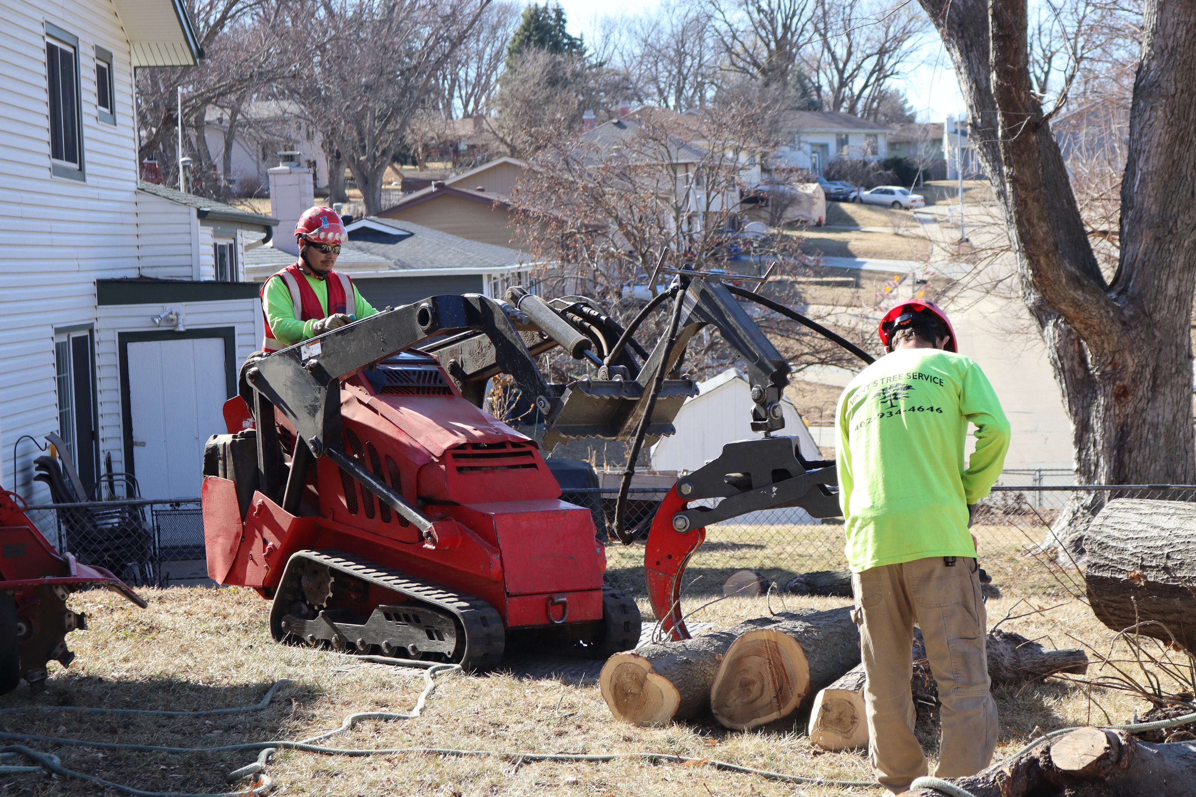Two Vinny's Tree Service crew members in green shirts operating skid steer to load large logs after tree removal
